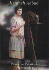 image shows an old photograph of the photographer herself stood by her camera (a box camera with a dark cloak on a tall tripod), perhaps retouched with a little colour but essentially sepia / black and white, with the text Karimeh Abbud, Pioneering Palestinian Photographer